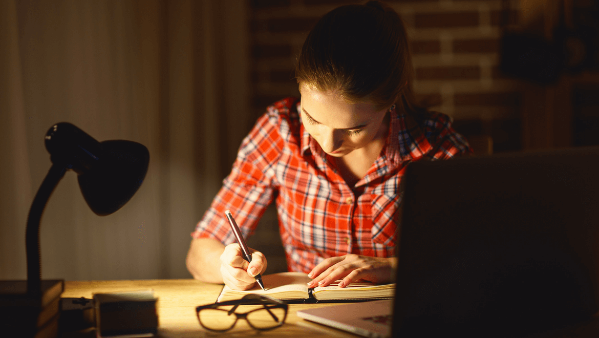 lighting and concentration: A woman with a red shirt at her desk writing with a desk light enlighting her project